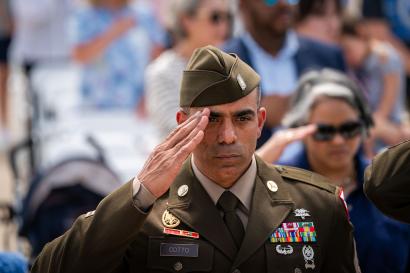Modern photograph of a US Army soldier in olive-colored dress uniform and cap standing outside in a crowd, saluting something behind the photographer.