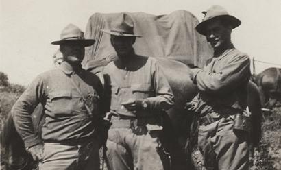 Black and white photograph of three unidentified soldiers posing for a picture. Center man is smoking a cigarette. In background is a covered wagon and horses. 