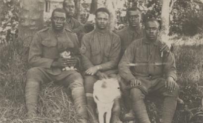 Black and white postcard photograph of five African American soldiers seated casually in wooded area with puppy on one soldier's lap and another dog in front center. 