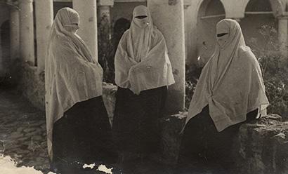 Black and white photograph of three people standing and sitting in a courtyard, dressed in skirts and veils covering their faces except for their eyes.