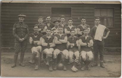 Black and white photograph of a group of men. The first row is sitting, and the rest are all standing, with the majority in football uniforms. One man stands to the side carrying a towel and another stands on the opposite side in uniform. There is a soccer ball at the front between the legs of one of the men.