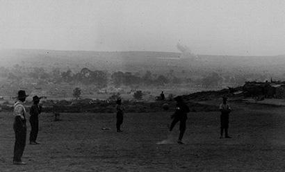 Black and white photo of a group of men in a field playing football (soccer). A bombshell explosion is going off in the distant background.