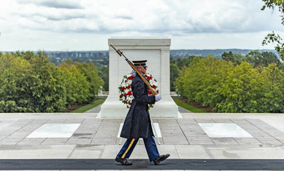 A white man in military uniform with a rifle on his shoulder marches in front of a rectangular white stone monument.