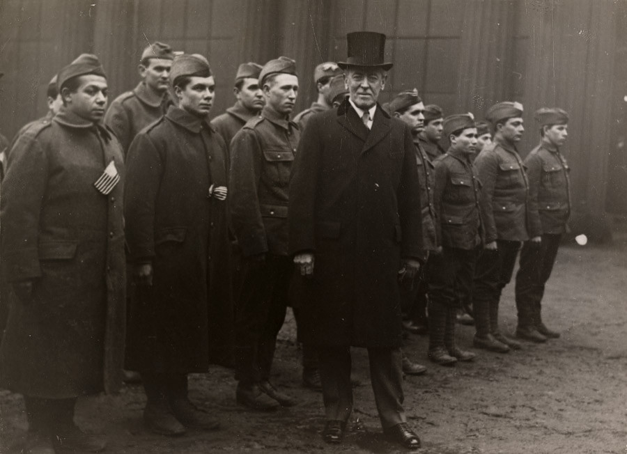 Photo en noir et blanc de Woodrow Wilson portant un haut-de-forme et un pardessus, debout devant un petit groupe de soldats alignés en uniforme militaire.