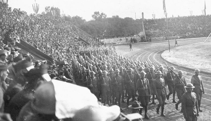 Photographie en noir et blanc de la piste autour d'un terrain de sport. Les tribunes sont remplies de spectateurs. Un régiment d'athlètes-soldats vêtus d'uniformes militaires avec des fusils sur leurs épaules marche sur la piste.