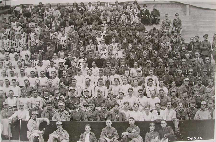 Photographie en noir et blanc d'une partie des tribunes d'un stade. Les personnes assises sont vêtues des uniformes sportifs de différents pays, regroupés par uniforme.
