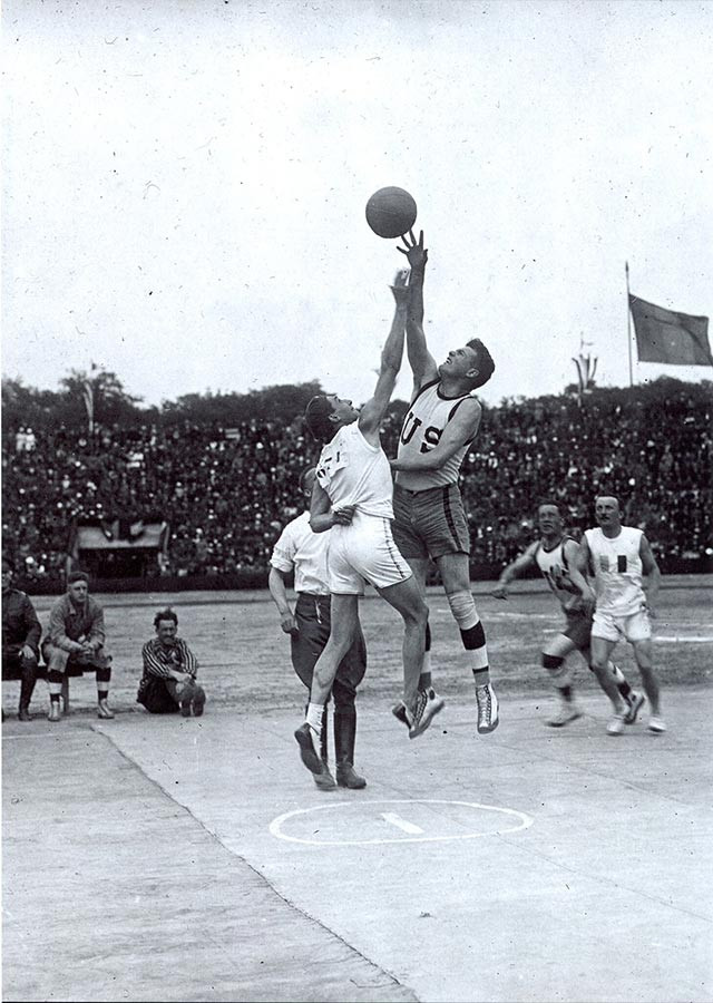 Photographie en noir et blanc d'un match de basket. Deux athlètes d'équipes adverses sautent dans les airs après le basket.