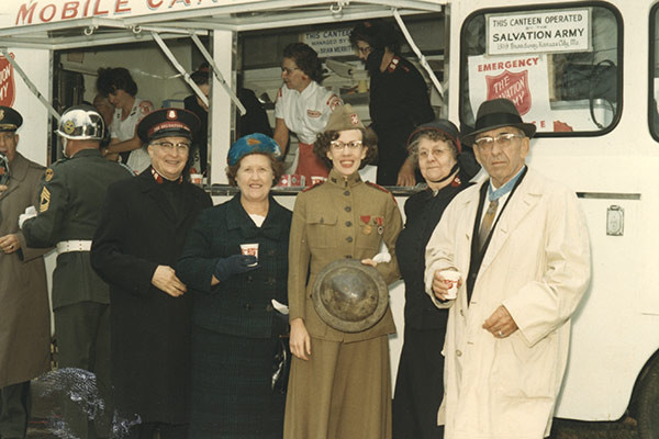 Salvation Army မိုဘိုင်းစားသောက်ဆိုင်ရှေ့တွင် WWI ခေတ်အဝတ်အစားနှင့် ယူနီဖောင်းများဝတ်ဆင်ထားသော လူအများအပြားဘေးတွင် ကတုတ်ကျင်းအင်္ကျီနှင့် ဦးထုပ်ကိုဝတ်ဆင်ထားသော အသက်ကြီးအဖြူရောင်အမျိုးသားတစ်ဦး၏ ခေတ်ဟောင်းအရောင်ဓာတ်ပုံ။
