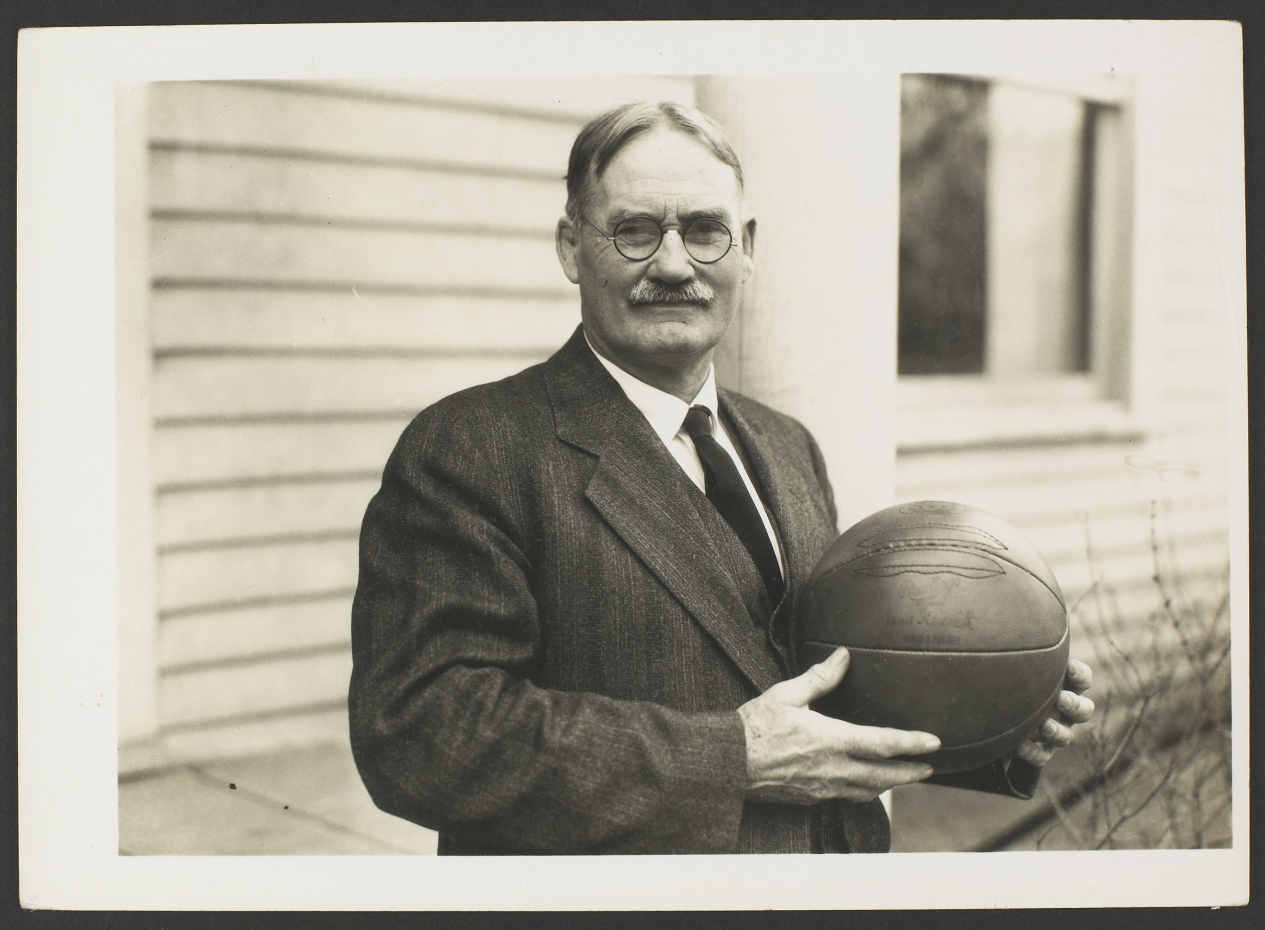 Photographie sépia d'un homme avec une moustache et des lunettes à monture métallique, vêtu d'un costume et tenant un ballon de basket