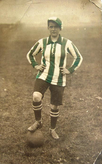 Photographie sépia d'une femme portant un maillot à rayures vertes et blanches, un pied en l'air sur un ballon de football.