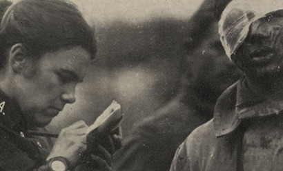 Black and white photograph of a woman in military uniform writing something down in a notebook standing next to a man in military uniform with a bloody bandage wrapped around his head and over one eye.