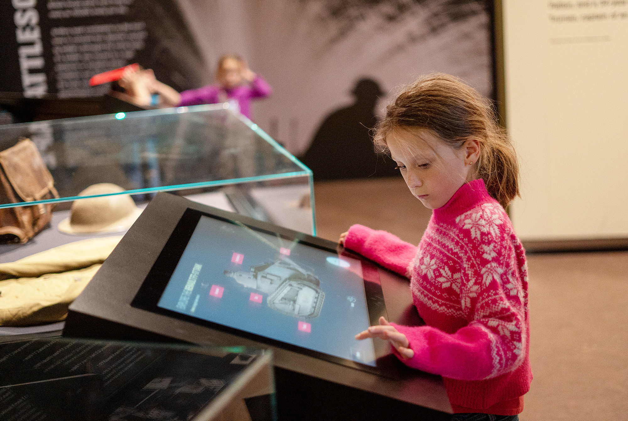 Modern photograph of a young white girl wearing a pink sweater about to press a button on an interactive display depicting a WWI tank