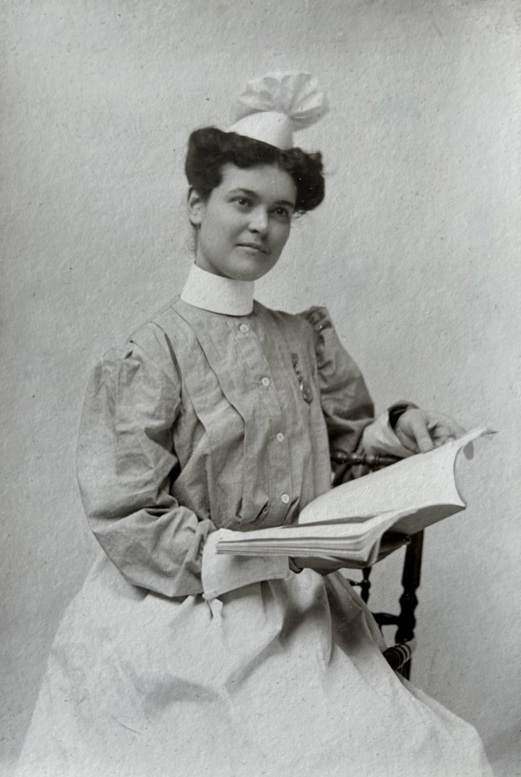 Black and white portrait photo of a white woman in an early 1900s nursing uniform and hat holding an open book