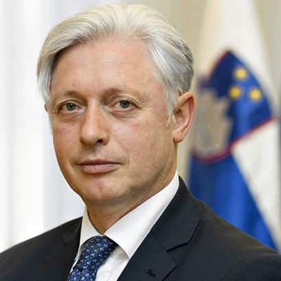Portrait photograph of a white man with white hair wearing a black suit and blue tie